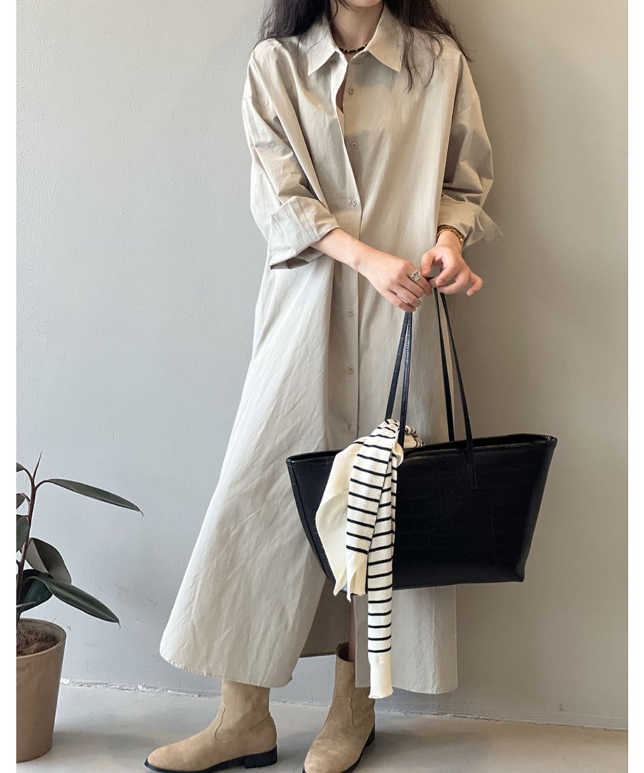 ベージュのロングシャツワンピースを着用した女性。ストライプ柄のニットをバッグに掛けて、茶色のブーツを履いている。