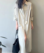 ベージュのロングシャツワンピースを着用した女性。ボタンを少し開け、ストライプ柄のニットを肩に掛け、黒いトートバッグを持っている。