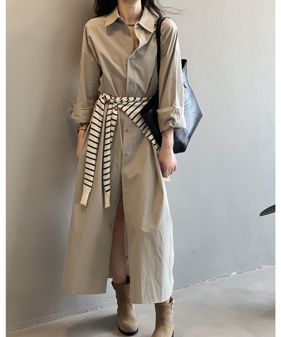 グレーのロングシャツワンピースを着用した女性が壁の近くに立っている。足元はベージュのスエードブーツを履いている。