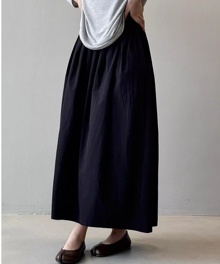 グレーの長袖Tシャツを着用し、ウエストから裾に向かってボリュームのある黒のロングスカートを着用したモデル。手はスカートのウエスト部分に添えられ、足元はブラウンの足袋型フラットシューズ。
