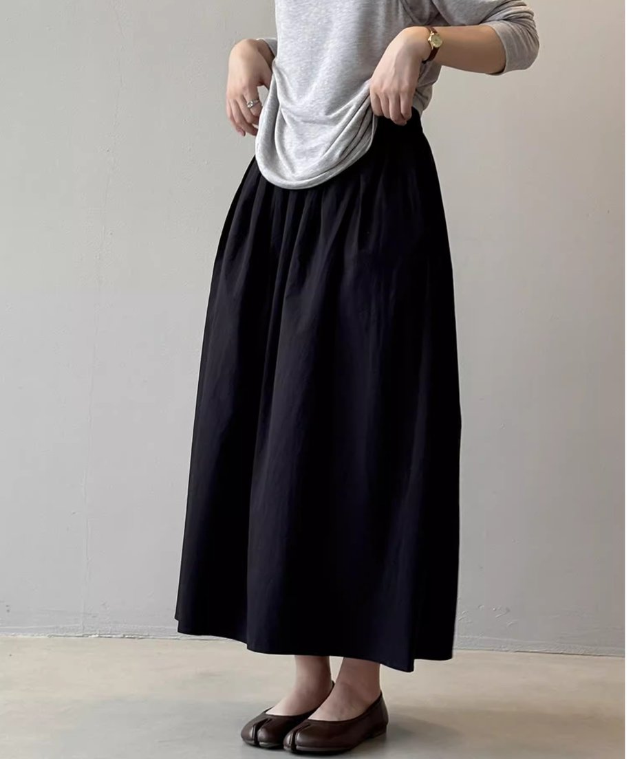 グレーの長袖Tシャツをウエストにたるませて着用したモデル。ウエストにギャザーとタックが入った黒のミモレ丈フレアスカートと、ブラウンの足袋型フラットシューズを着用。