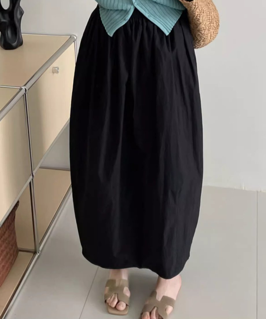 黒色のロングバドスカートを着用した女性の下半身。水色のカーディガンを合わせ、かごバッグを持っている。