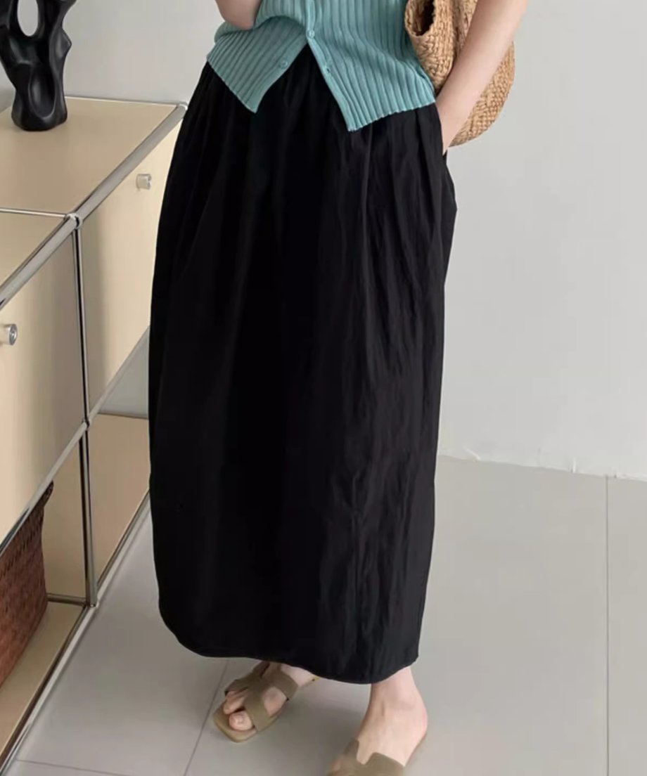 黒色のロングバドスカートを着用した女性の下半身。水色のカーディガンを合わせ、かごバッグを持っている。