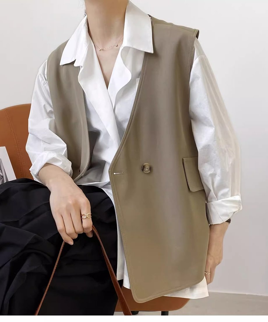 ベージュのダブルブレスト風ジレベストを、白の長袖シャツと黒のボトムスに重ね着した女性が椅子に座ったショット。大人のきれいめな上半身ファッション。