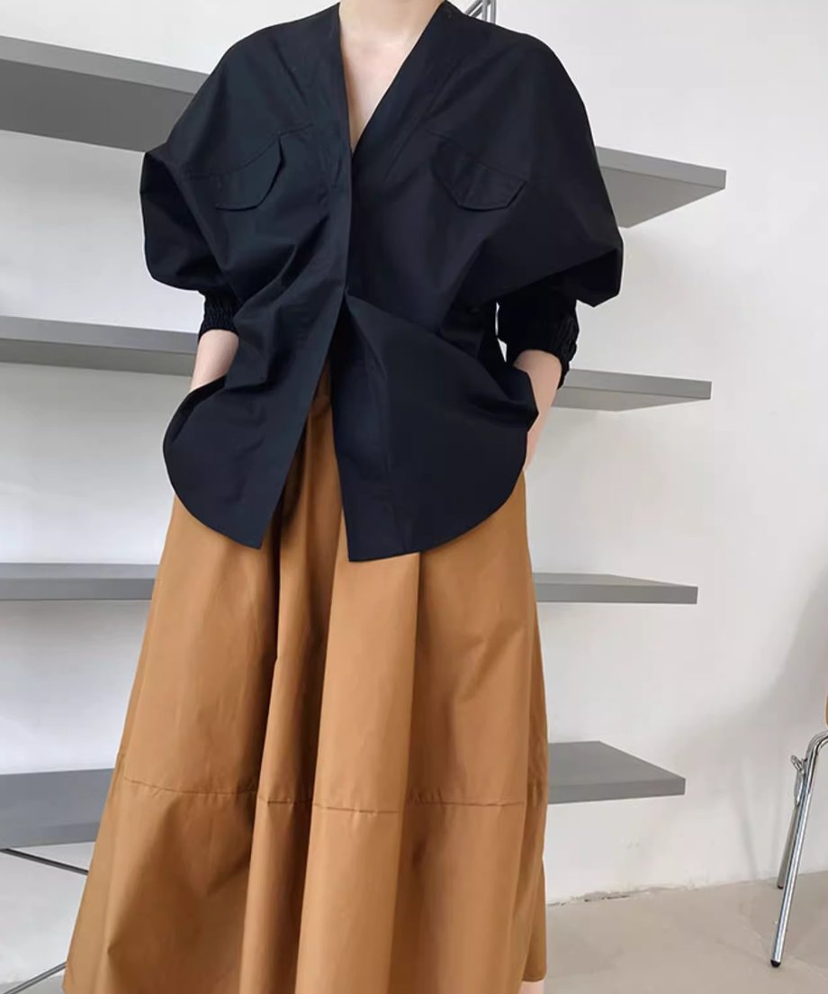 Vネックとドルマンスリーブの黒いシャツを、茶色のフレアスカートと合わせて着用し、手をポケットに入れている女性のウエストアップショット