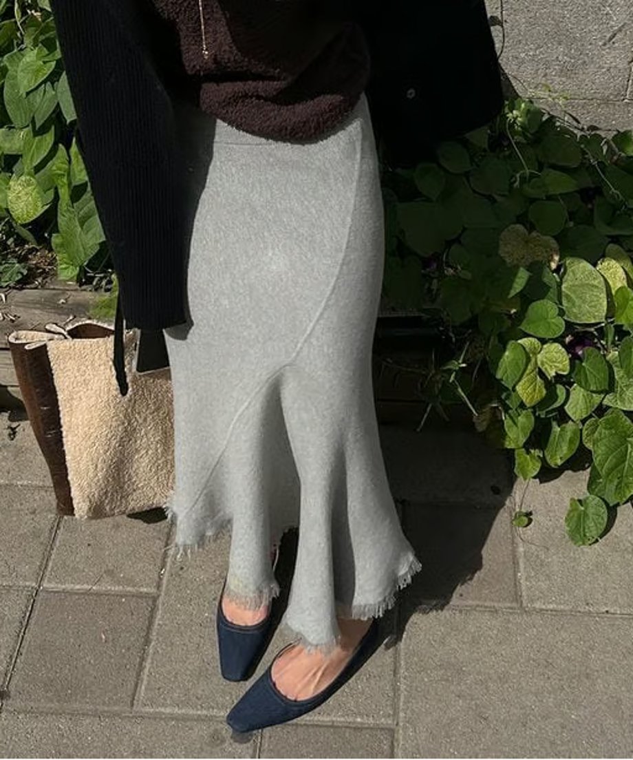 ライトグレーのニットマーメイドスカート。裾にフリンジ加工が施されており、黒いカーディガンと合わせて着用している女性の屋外ショット。足元にはブラウン系のバッグが見える。
