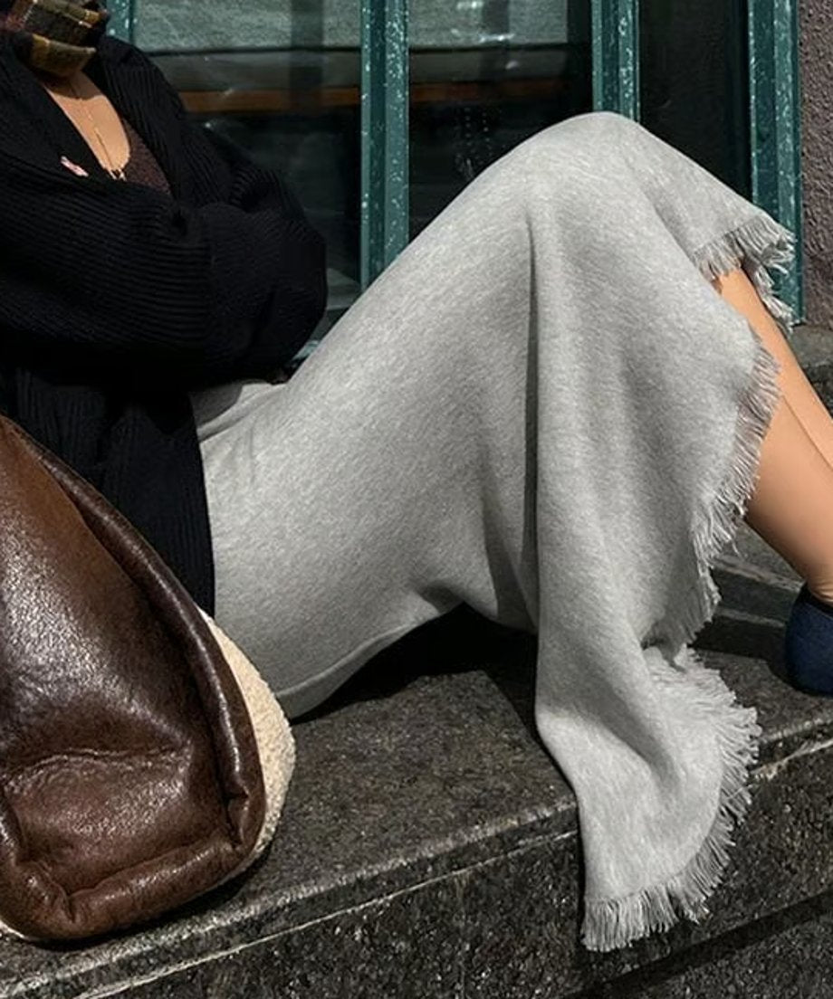 ライトグレーのニットマーメイドスカート。裾にフリンジ加工が施されており、黒いトップスと合わせて着用している女性が屋外で座っている画像。