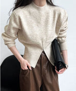 ベージュのハイネックニットプルオーバー。裾にジッパー付きの深いフロントスリットがあり、ブラウンのレザー調パンツと黒いバッグを合わせた女性。