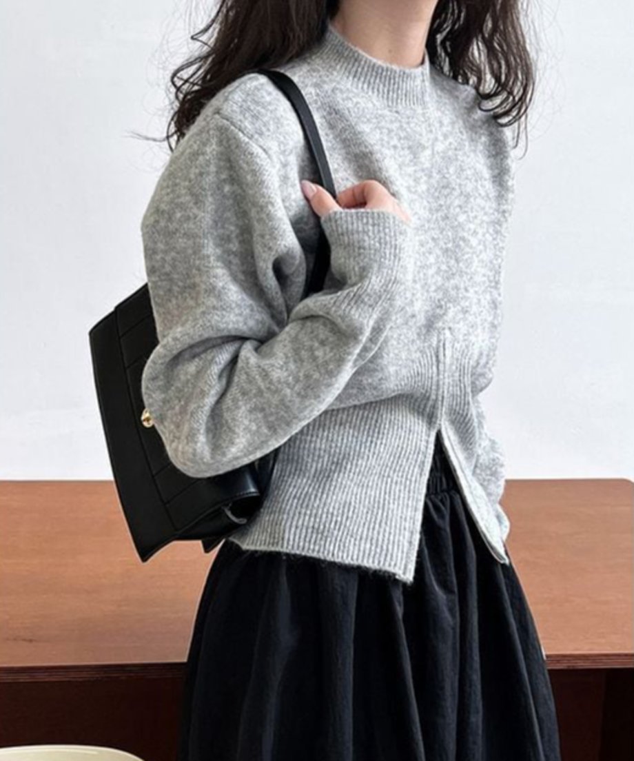 グレーのハイネックニットプルオーバー。裾にジッパー付きの深いフロントスリットがあり、黒いロングスカートと合わせて着用している女性の上半身。黒いバッグを肩にかけている。