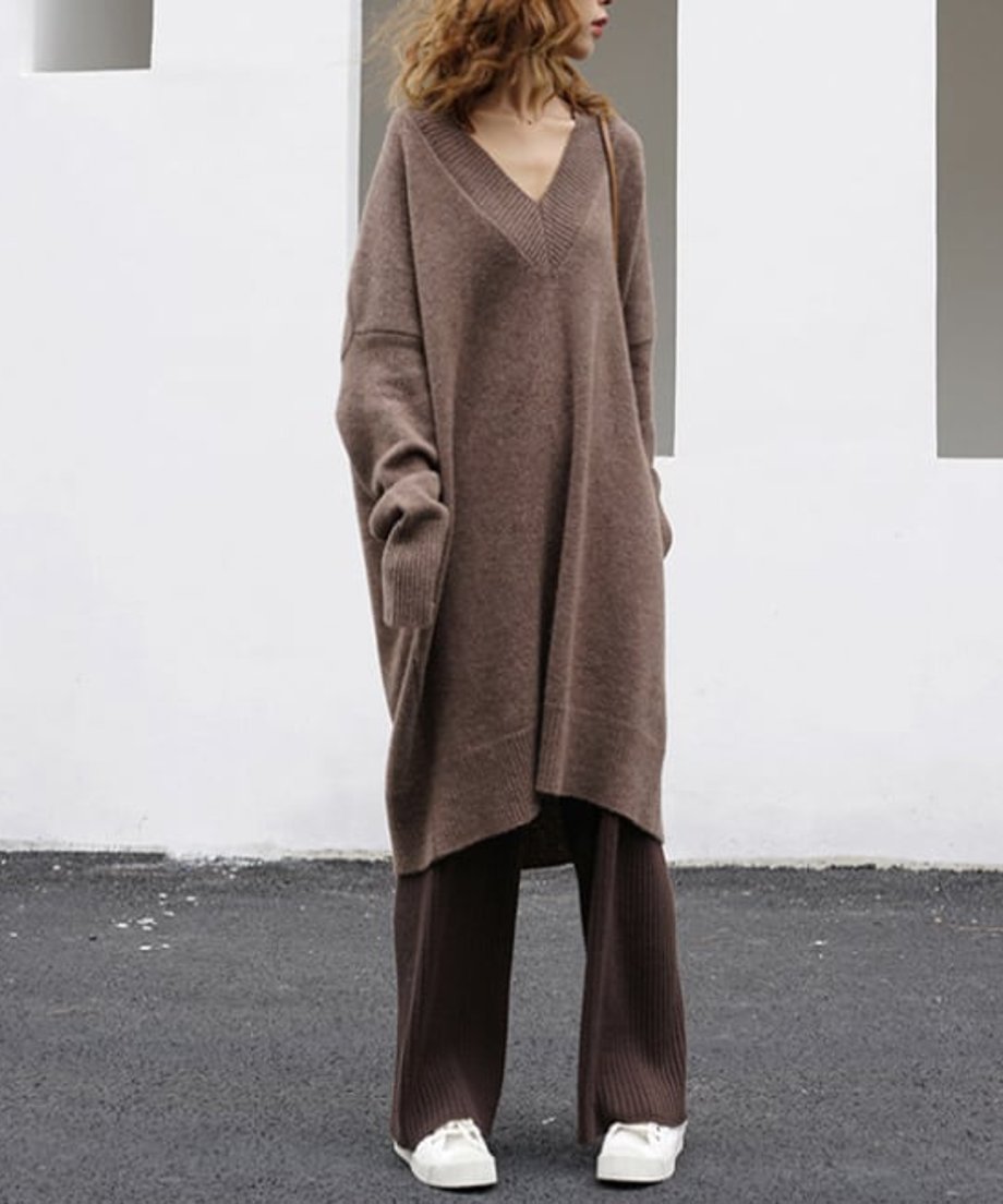 キャメルのVネックニットワンピースを着用した女性がブラウンのパンツと白いスニーカーを合わせたコーディネート。屋外の白い壁の前に立っている。