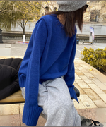 ブルーのクルーネックニットプルオーバー。ライトグレーのスウェットパンツと合わせてベンチに座る女性の上半身。