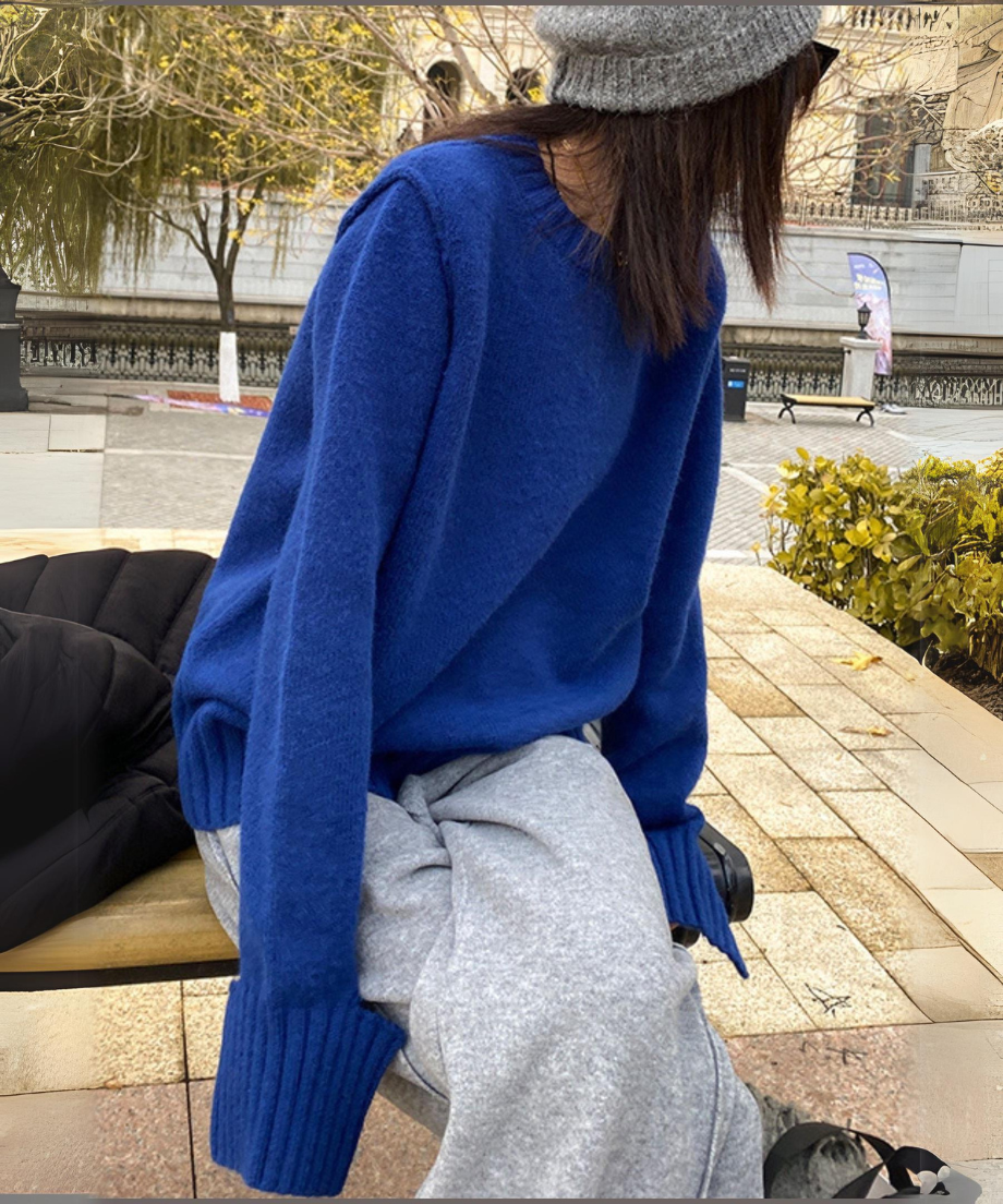 ブルーのクルーネックニットプルオーバー。ライトグレーのスウェットパンツと合わせてベンチに座る女性の上半身。