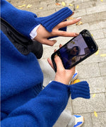 ブルーのクルーネックニットプルオーバー。幅広リブの袖口が特徴的。スマホを手にして自分に向けている。