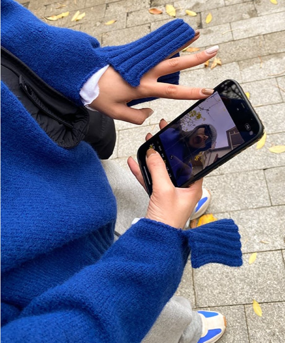 ブルーのクルーネックニットプルオーバー。幅広リブの袖口が特徴的。スマホを手にして自分に向けている。