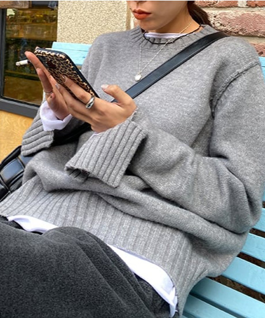 グレーのクルーネックニットプルオーバー。幅広リブの袖口が特徴的で、ダークグレーのボトムスと合わせ、ブルーのベンチに座る女性の上半身。