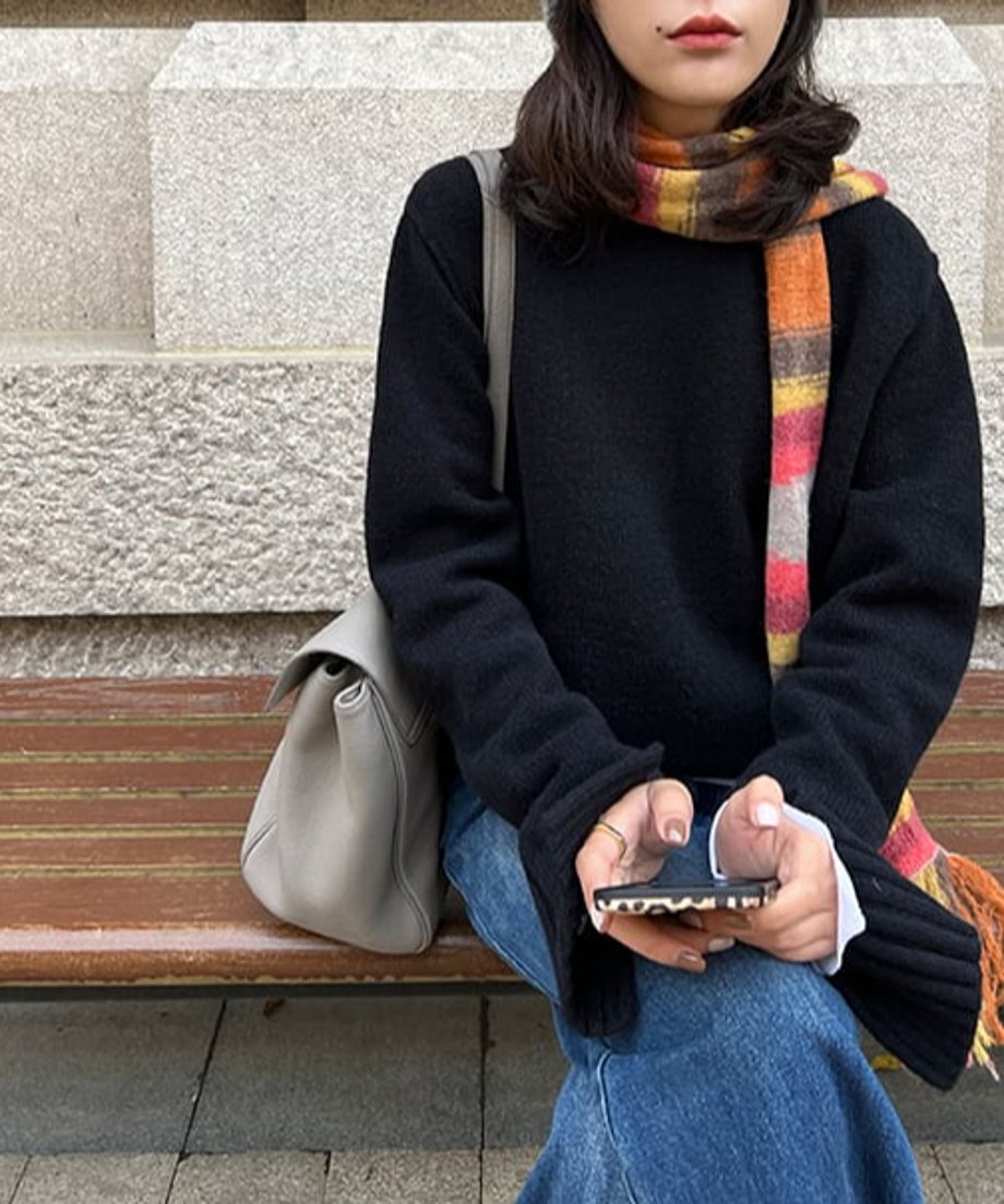 黒のクルーネックニットプルオーバー。ライトグレーのバッグとデニムと合わせて着用し、カラフルなマフラーを巻いた女性