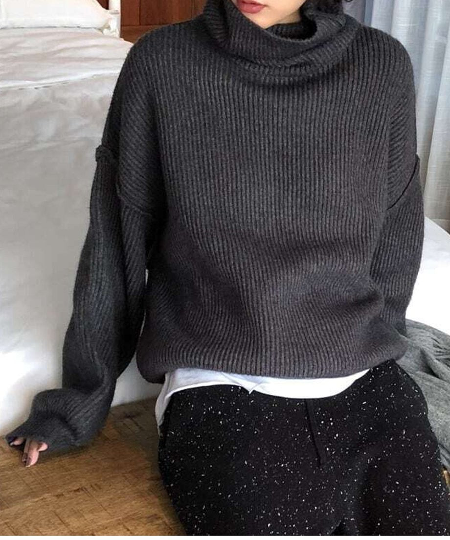 グレーのゆるっとしたハイネックニットを着用し、椅子に深く座っている女性の画像。