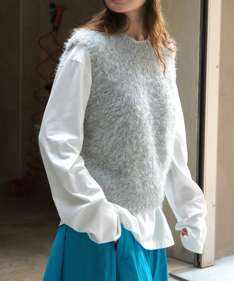 グレーの毛足の長いニットベストと白のロングTシャツ、鮮やかなブルーのロングスカートを着用した女性のやや斜めからのショット。白い太めのスリーブのシャツを合わせている。