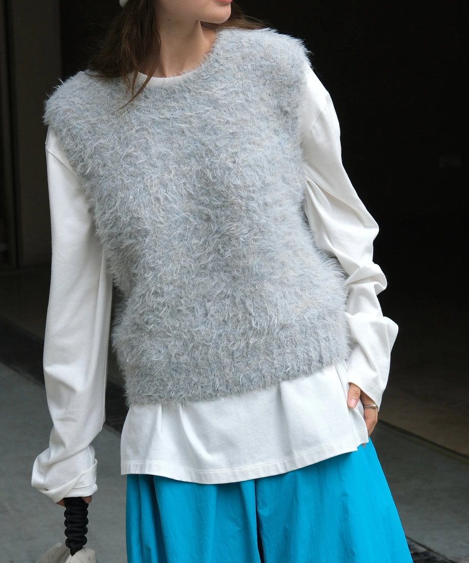 グレーの毛足の長いニットベストと白のロングTシャツ、鮮やかなブルーのロングスカートを着用した女性の正面画像。