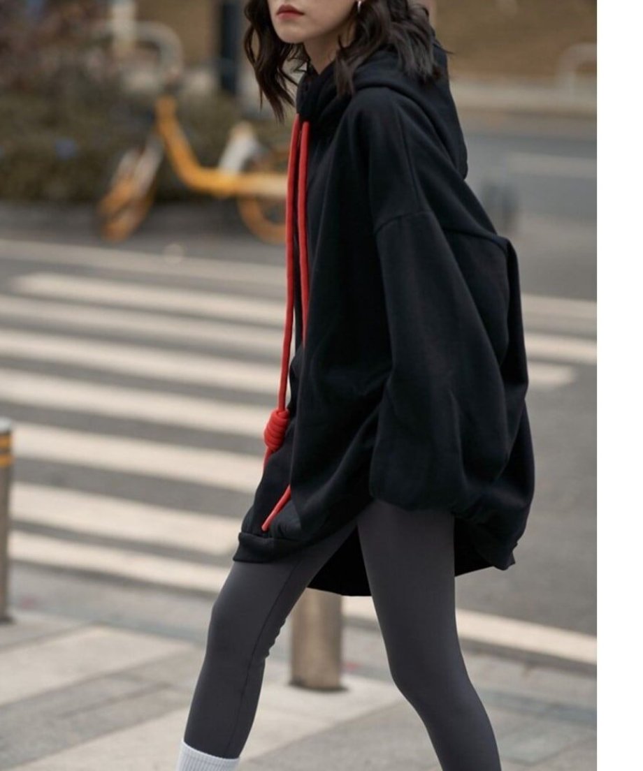 黒のオーバーサイズロングパーカーを着用した女性が、横断歩道付近を横向きに歩いている全身の様子。パーカーの裾が斜めにカットされたデザインと灰色のレギンス、白いソックスが確認できる。