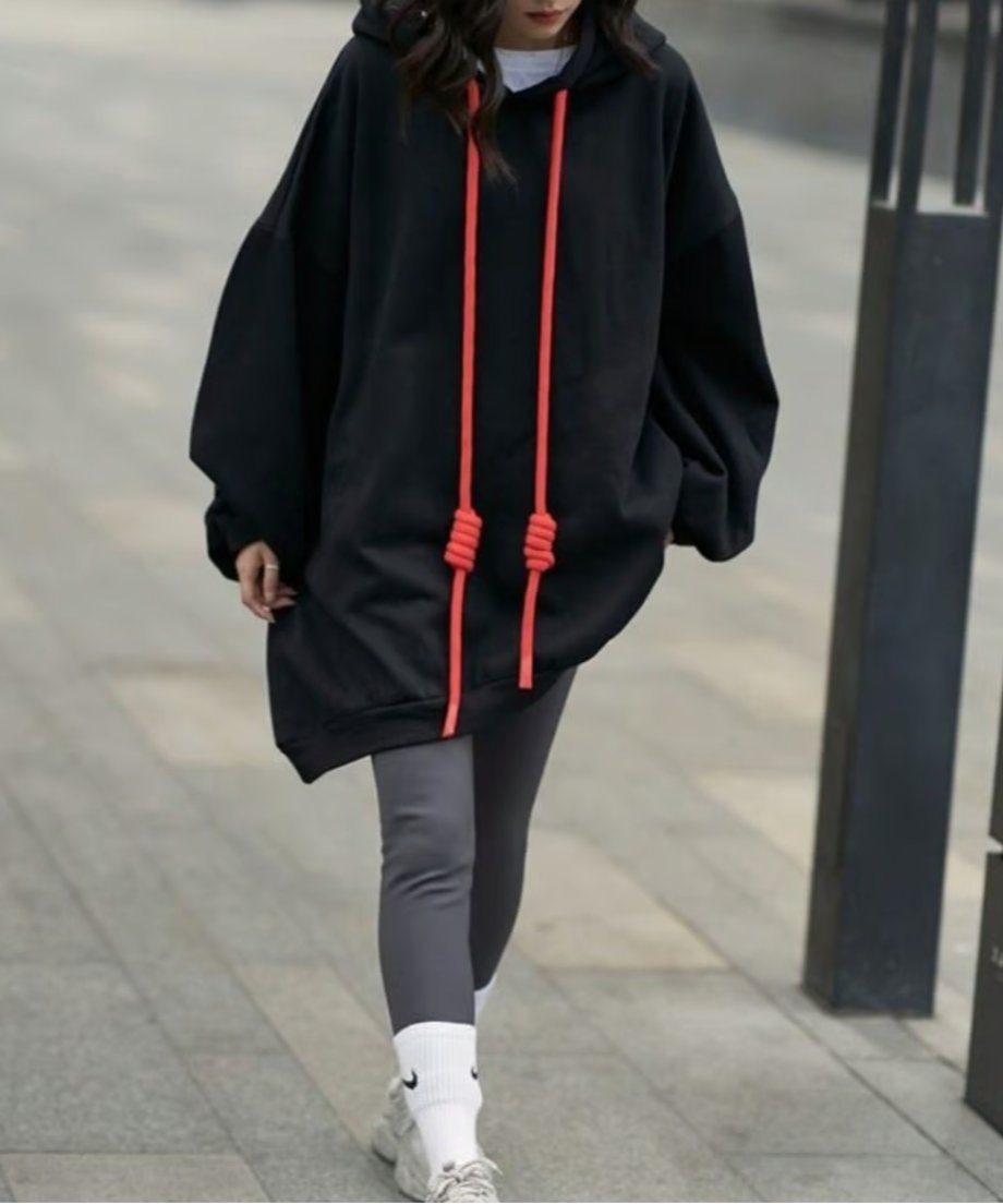 黒のオーバーサイズロングパーカーを着用し、灰色のレギンスと白いスニーカーを合わせた女性が、街中の歩道を歩いている全身の様子。パーカーのドローコードの結び目が確認できる。
