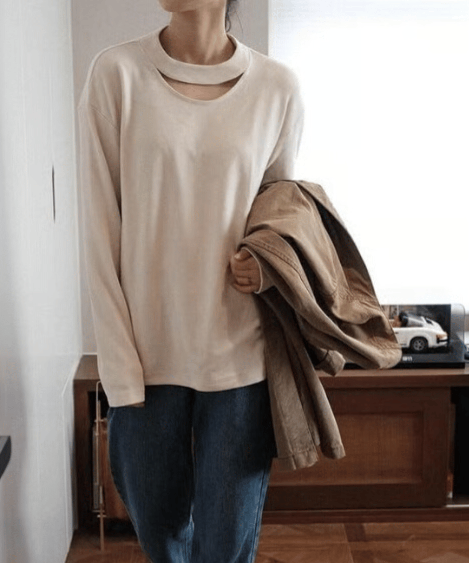 ライトアプリコットの長袖スリットネックトップスを濃いデニムパンツに合わせ、茶系のジャケットを手に持った全身ファッション画像。