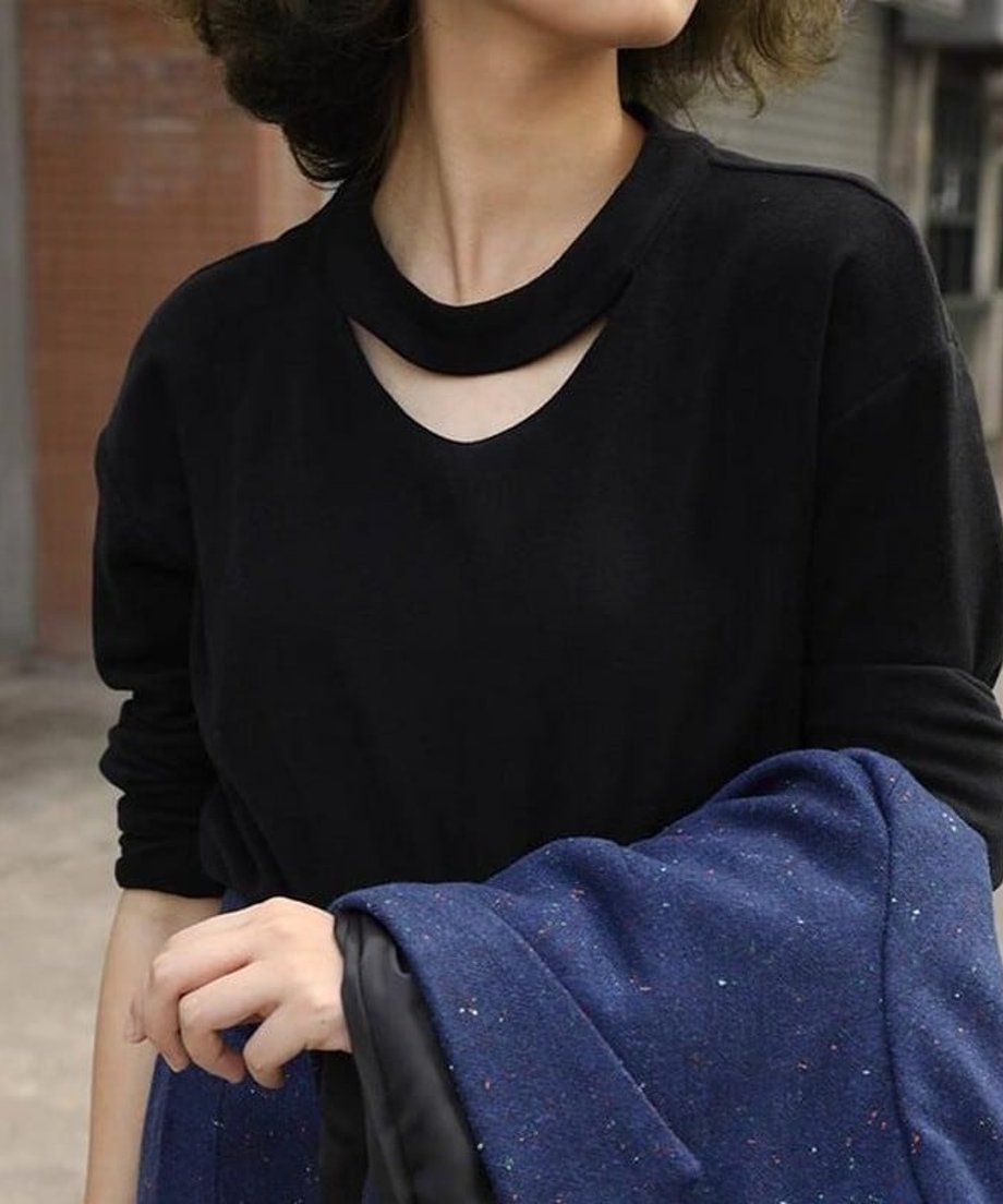 ブラックの長袖スリットネックトップスを着用した女性が手にブルー系のネップ入りのジャケットを持ったバストアップショット。