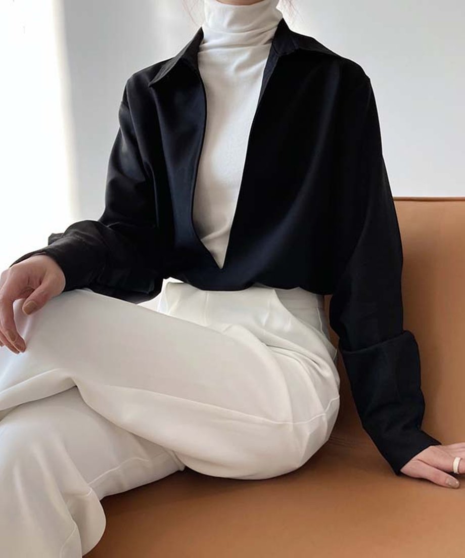 白いタートルネックの上に黒のディープVネックシャツを重ね着し、袖をロールアップした女性が、白いハイウエストパンツを穿いてソファに座っている上半身のコーディネート。