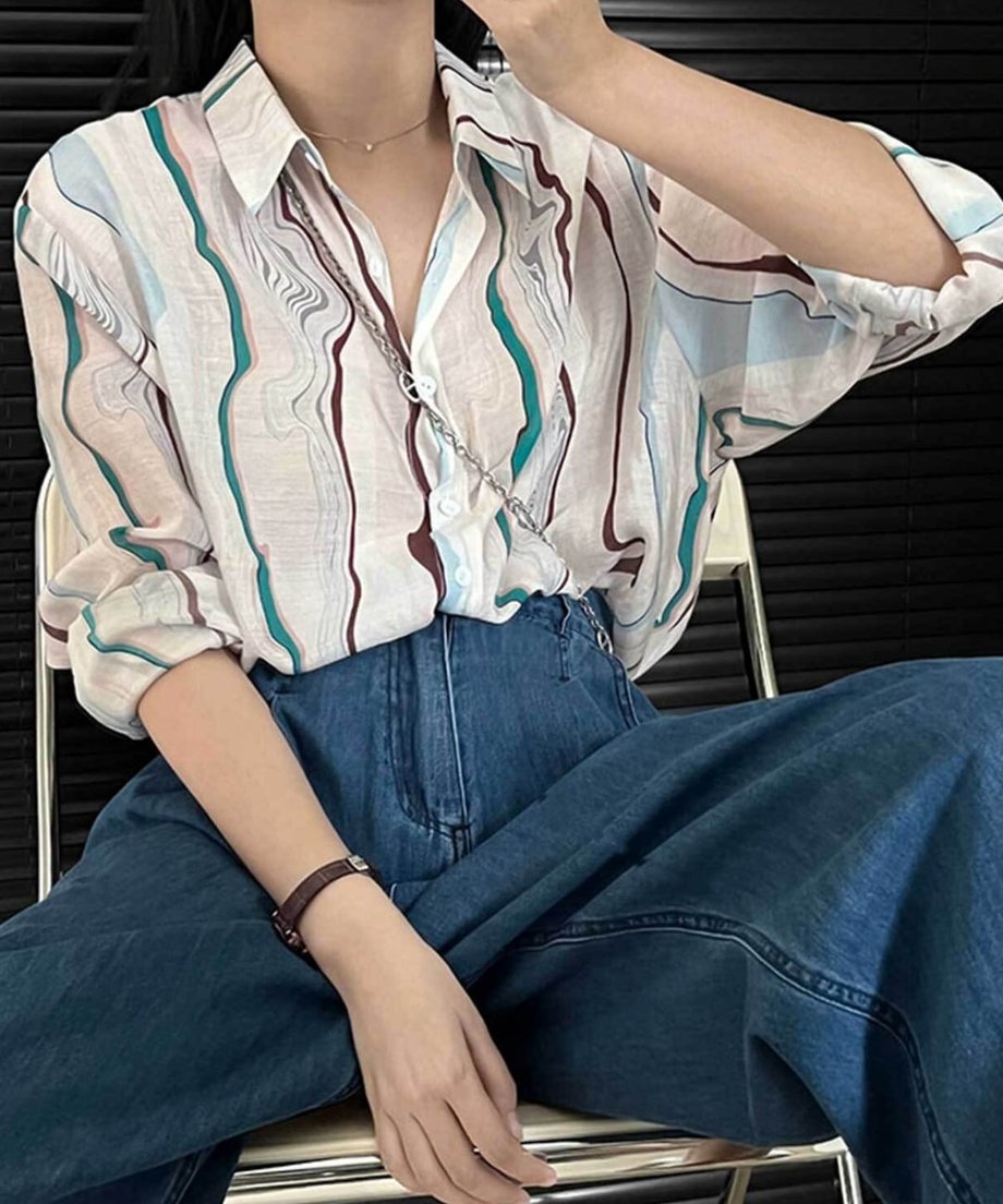 マーブル柄シアーシャツをデニムのワイドパンツと合わせ、椅子に座っている女性の全身像。