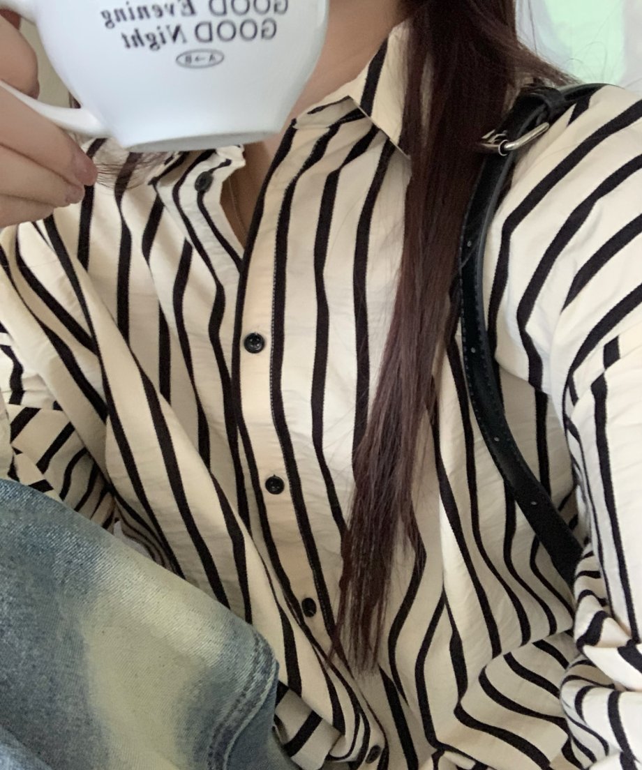 オフホワイトに黒のストライプが入った長袖シャツを着た女性のクローズアップ。コーヒーカップを持ち、デニムを着用。