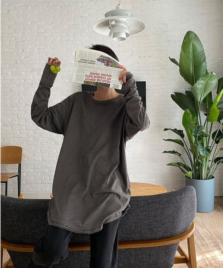 グレーのロングスリーブTシャツを着用した女性の室内ショット。手には新聞を持っている。