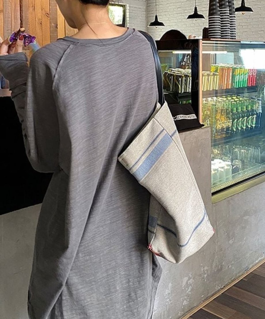 グレーのロングスリーブTシャツを着用し、ベージュと青のストライプ柄トートバッグを肩にかけた女性の後ろ姿。