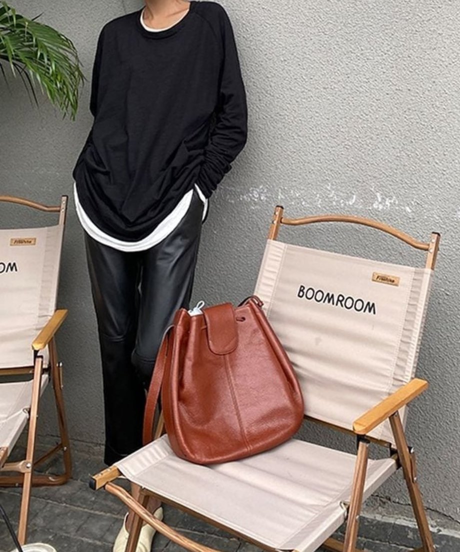 ロングスリーブTシャツに、白のインナーを重ね、黒のレザー調パンツを着用した女性の全身ショット。椅子にブラウンのバッグが置かれている。
