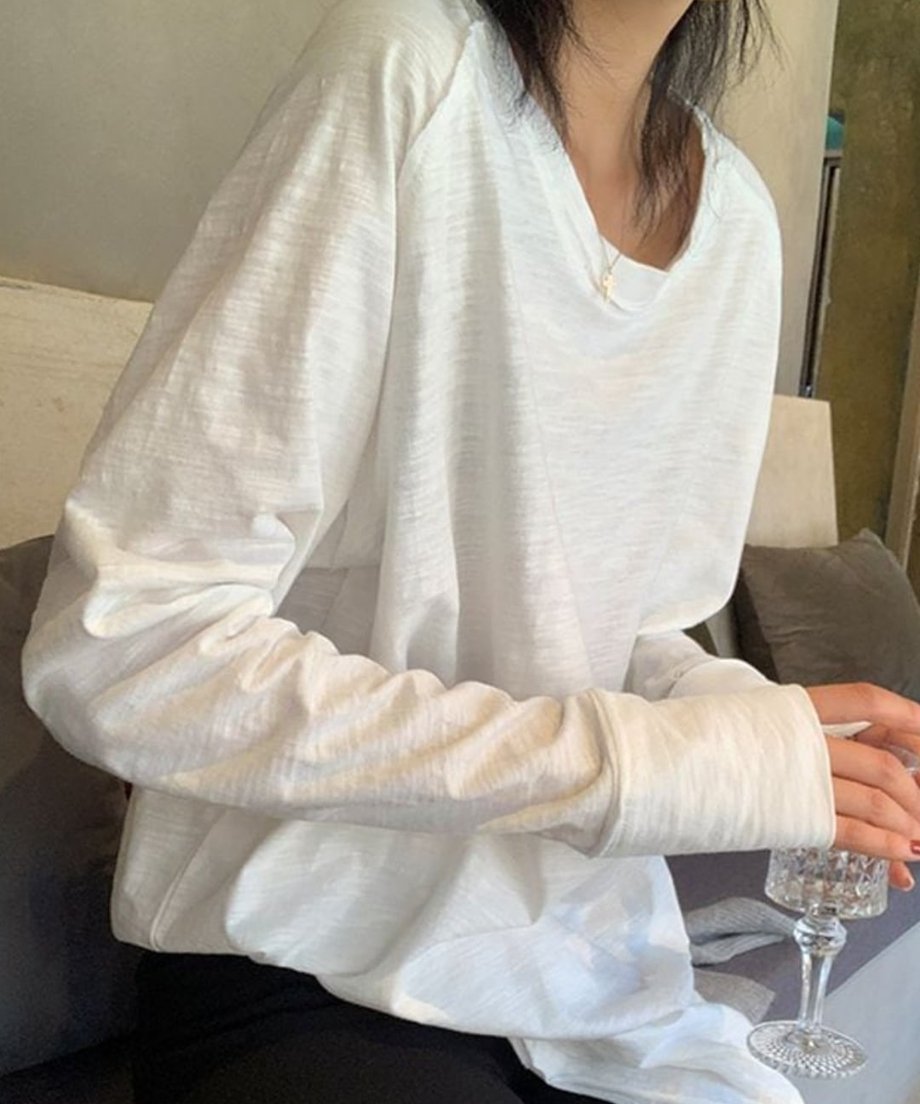 ホワイトのロングスリーブTシャツを着用し、椅子に座っている女性のバストアップ。デコルテ周りが見える緩やかなネックライン。