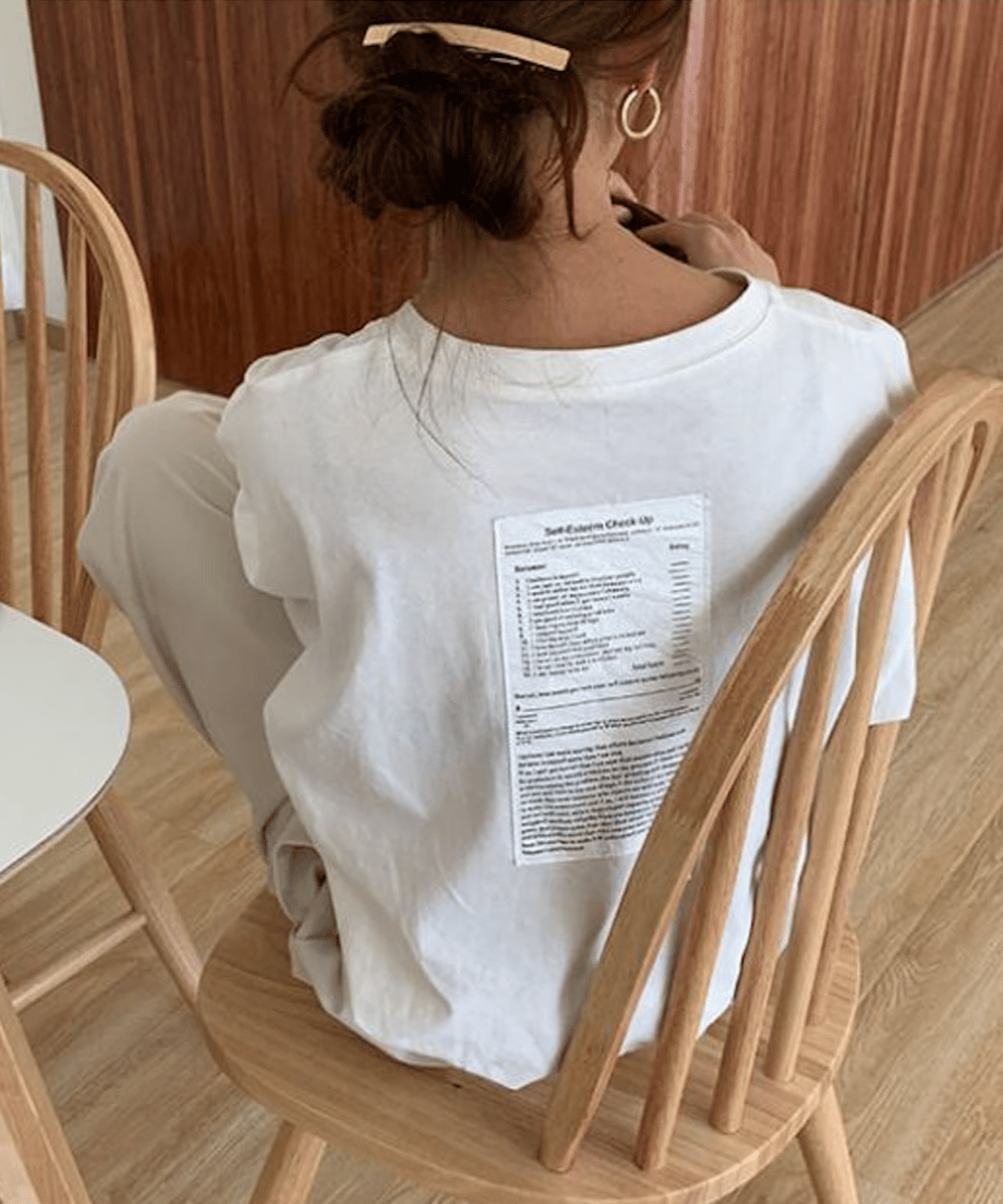 白のオーバーサイズ長袖Tシャツを着た女性が白い椅子に座っている後ろ姿。背中には縦長のテキストプリント（ケアラベル風）があり、黒いニットの斜めがけバッグを着用。