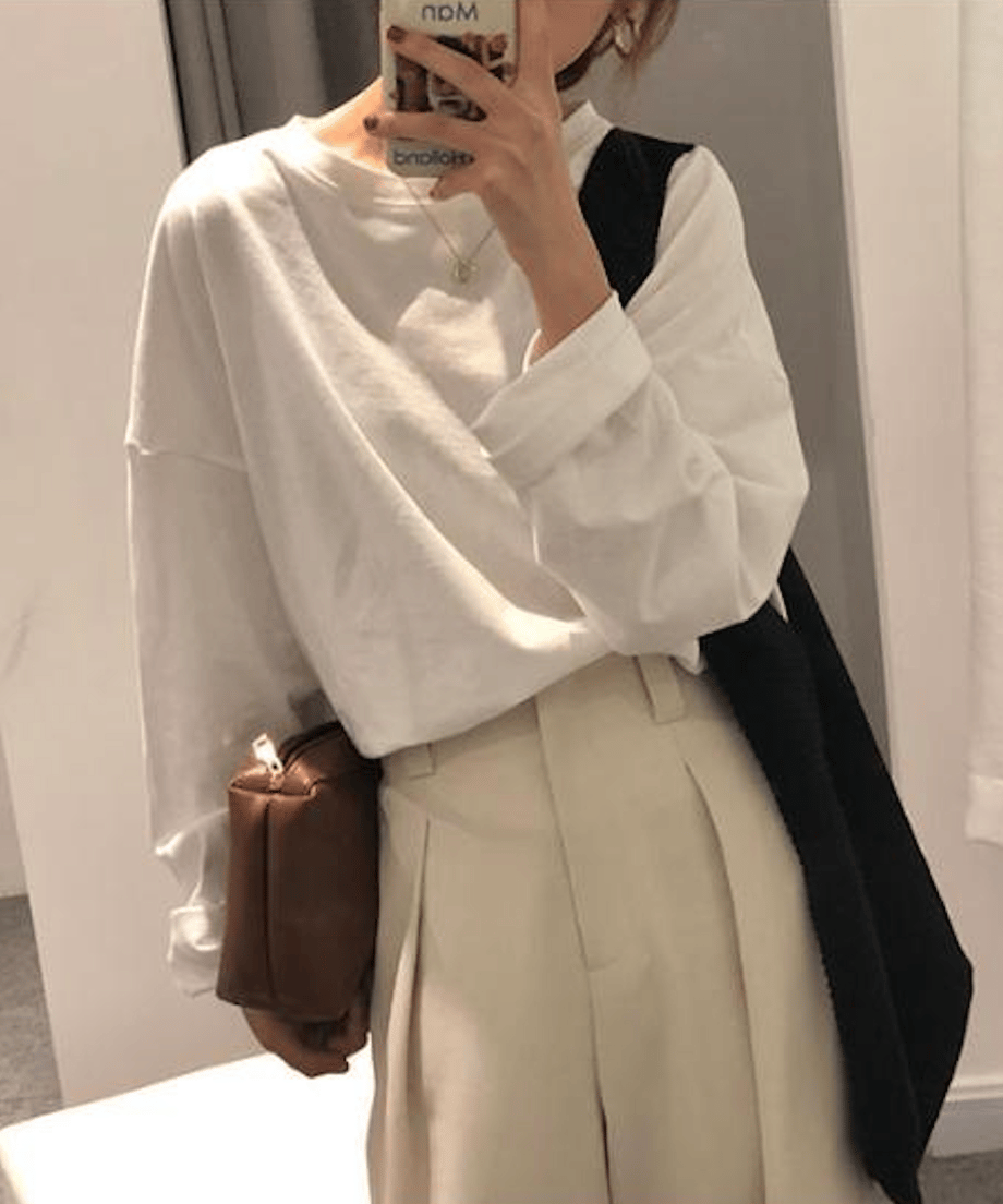 白のオーバーサイズ長袖Tシャツを着用し、スマートフォンを持ってポーズをとる女性。ベージュ系のタック入りワイドパンツ、黒い斜めがけバッグ、ブラウンのミニバッグを合わせている。