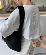 白のオーバーサイズ長袖Tシャツを着た女性の背中側斜めからのクローズアップ。白い縦長のテキストプリント（ケアラベル風）が確認でき、黒いニットの斜めがけバッグを着用。