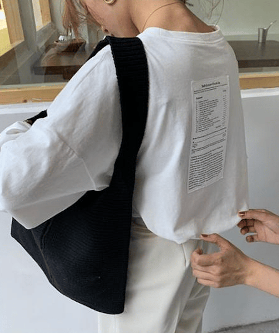 白のオーバーサイズ長袖Tシャツを着た女性の背中側斜めからのクローズアップ。白い縦長のテキストプリント（ケアラベル風）が確認でき、黒いニットの斜めがけバッグを着用。
