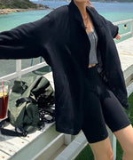 黒のシアー素材テーラードジャケットを着た女性は屋外の白いベンチの前で大きく手を広げたショット。