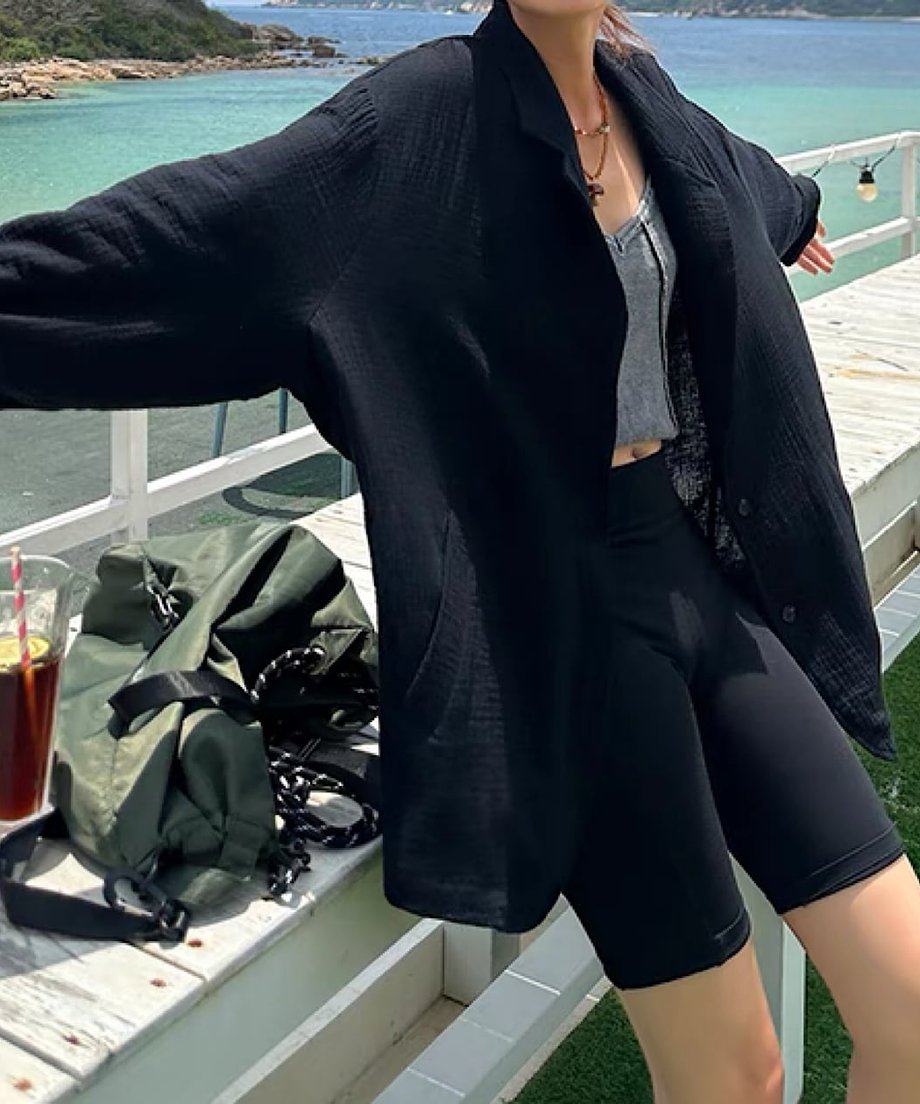 黒のシアー素材テーラードジャケットを着た女性は屋外の白いベンチの前で大きく手を広げたショット。