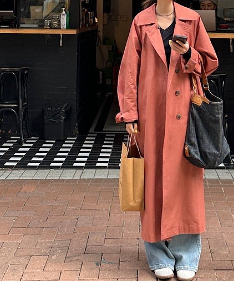 ダスティレッドのロングトレンチコートを着た女性が屋外に立っているショット。大きなバッグと紙袋を持ってスマホを見ている。