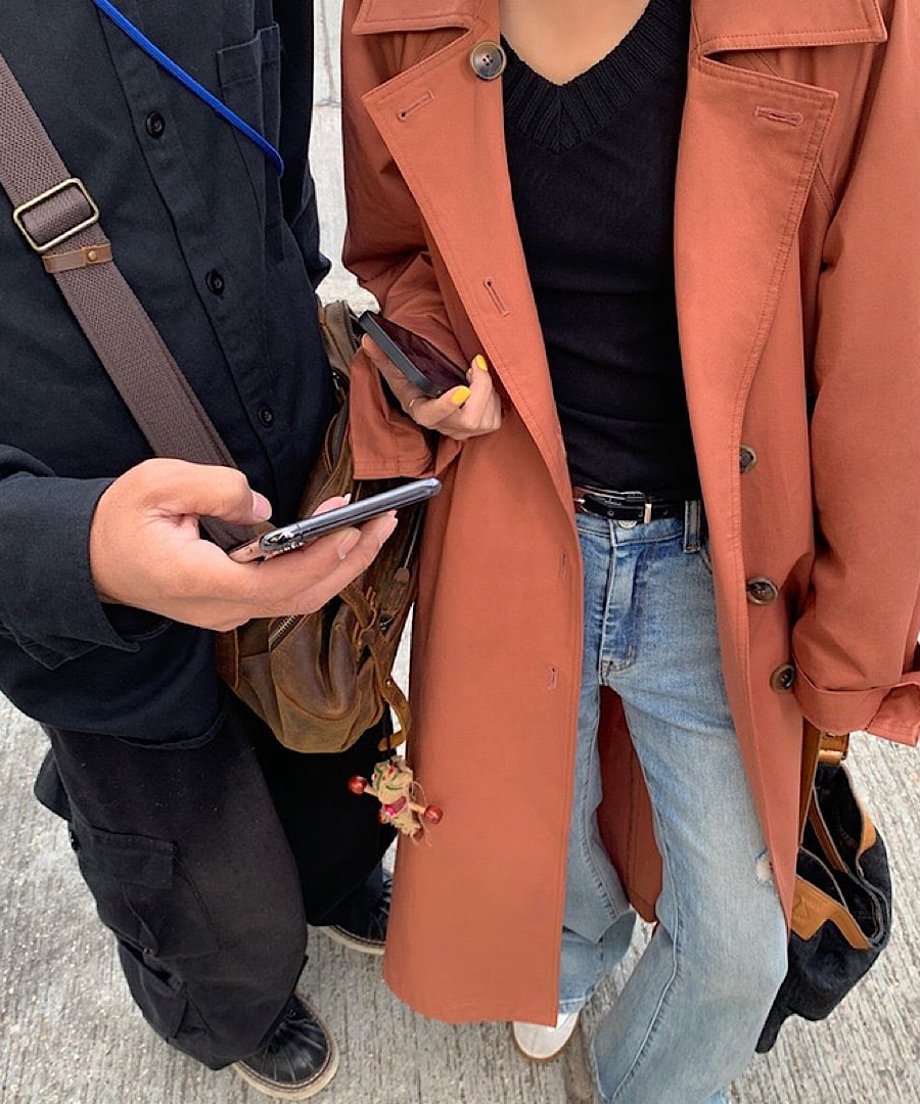 ダスティレッドのロングトレンチコートを着た女性と黒いアウターを着た男性が並んで立っている全身像。トレンチコートの女性は青いデニムパンツを着用している。