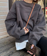 グレーのオーバーサイズスウェットを着用し、木製のベンチに座っている女性。茶色のショルダーバッグを斜めがけし、スマートフォンを持っています。