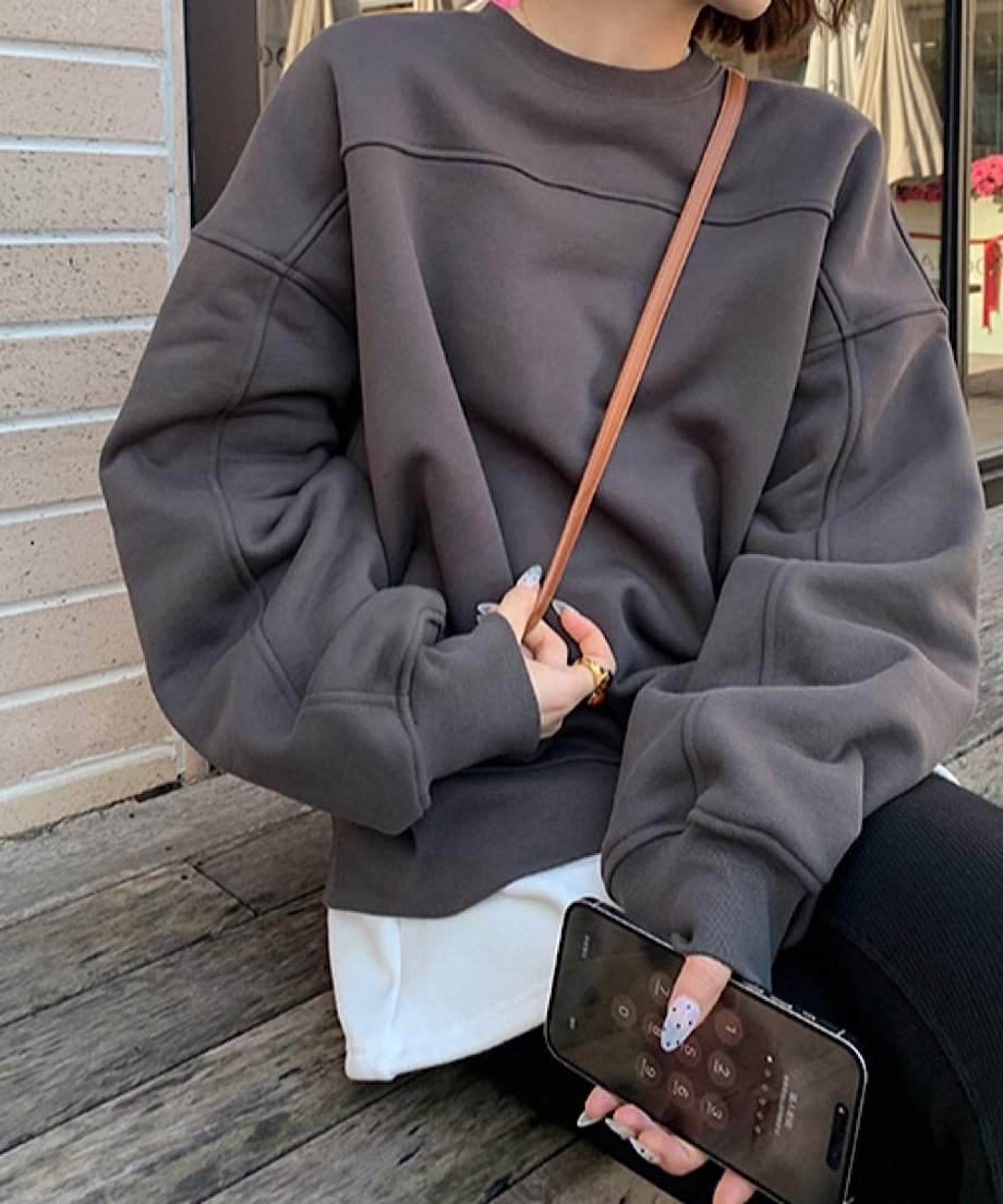 グレーのオーバーサイズスウェットを着用し、木製のベンチに座っている女性。茶色のショルダーバッグを斜めがけし、スマートフォンを持っています。