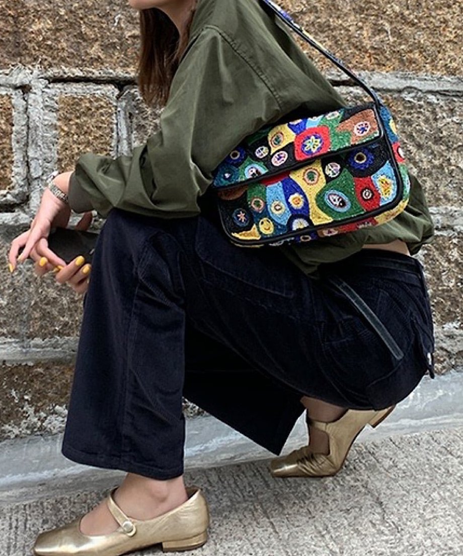 ミリタリーグリーンのプルオーバーと黒のワイドパンツ、ゴールドのシューズを履いてしゃがんでいる女性。カラフルなビーズバッグを着用。