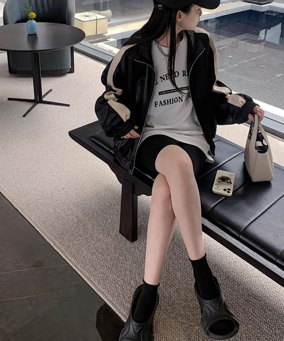 黒のブルゾンを羽織り、白のロゴTシャツと黒のサイクリングショーツを着用した女性が、室内のベンチに座っている。黒のキャップと厚底サンダルを合わせ、白のミニバッグを持っている。