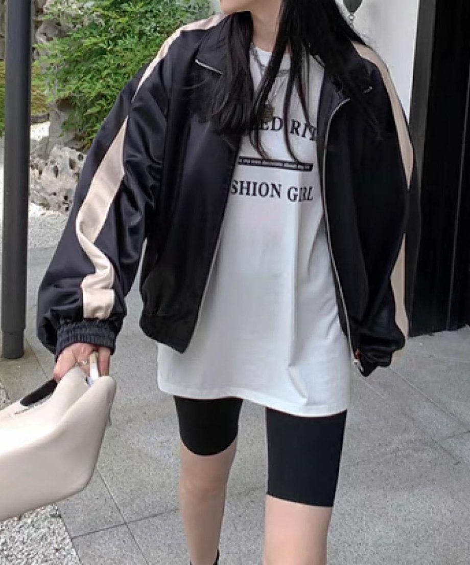 黒のブルゾンを着用した女性の屋外での上半身ショット。インナーに白のロゴTシャツ、ボトムスに黒のサイクリングショーツを合わせ、白のバッグを持っている。
