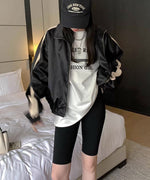 黒のブルゾンを羽織り、白のオーバーサイズTシャツと黒のサイクリングショーツを着用した女性の全身像。黒のキャップを被り、ベッドの横でポーズをとっている。