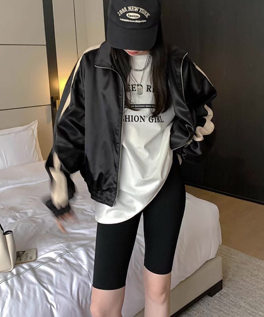 黒のブルゾンを羽織り、白のオーバーサイズTシャツと黒のサイクリングショーツを着用した女性の全身像。黒のキャップを被り、ベッドの横でポーズをとっている。