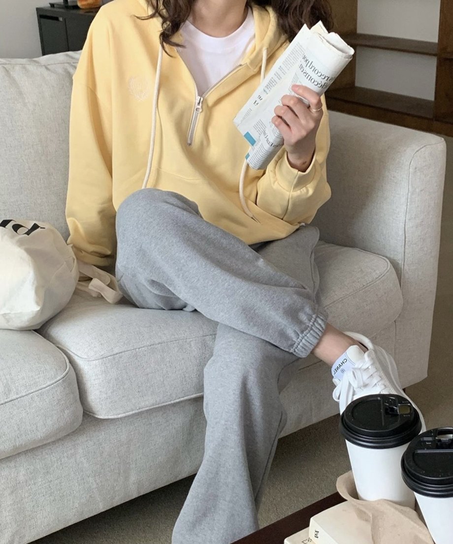 イエローのハーフジップパーカーを着用した女性が、グレーのスウェットパンツと白いロゴ入りトートバッグを合わせ、ソファで新聞を持っている。